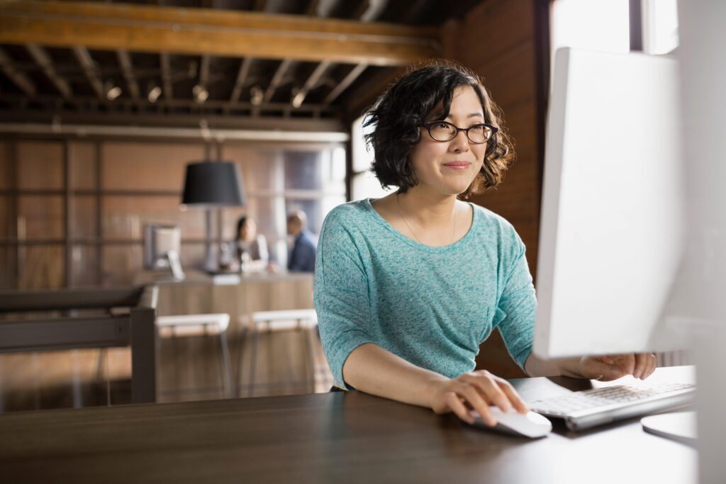 A person wearing glasses and a teal top sits at a desk, using a mouse and keyboard at a desktop computer in a warmly lit open office.