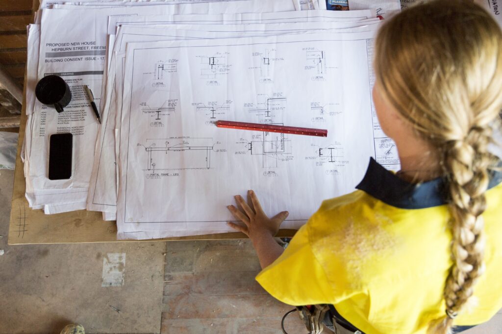 A construction worker reviews structural drawings on-site, studying detailed framing and connection plans for a new house build.