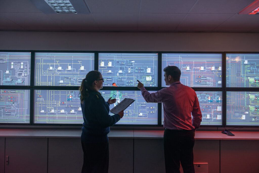 Two operators review system schematics in a control room, discussing readings across a wall of monitors displaying piping, fuel, and cooling diagrams.