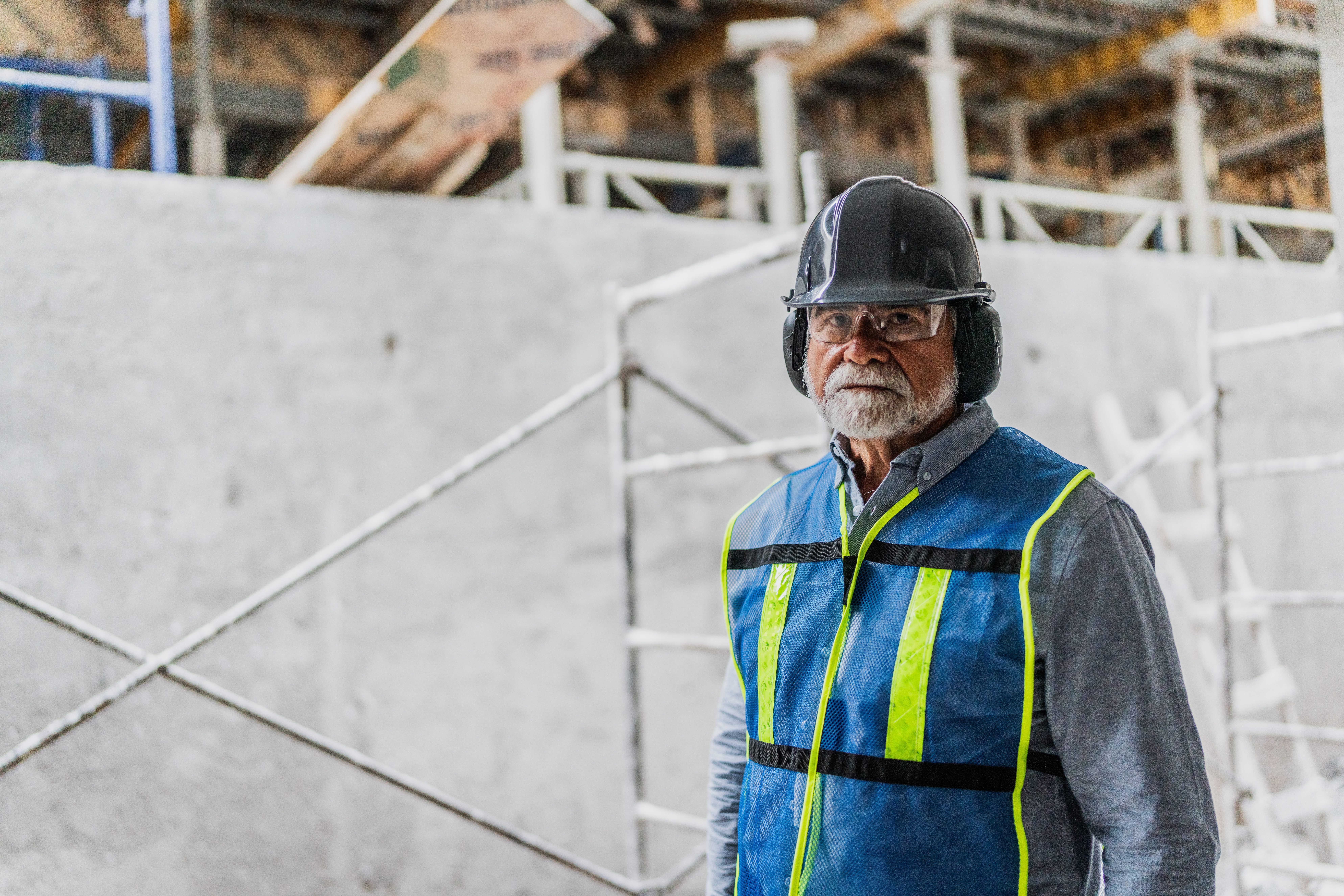 A bearded older worker wearing a black hard hat, ear protection, clear safety glasses, a gray button-down shirt, and a blue high-visibility safety vest with neon yellow stripes stands on a construction site. Concrete walls, white scaffolding poles, and wooden beams are visible in the soft-focus background.