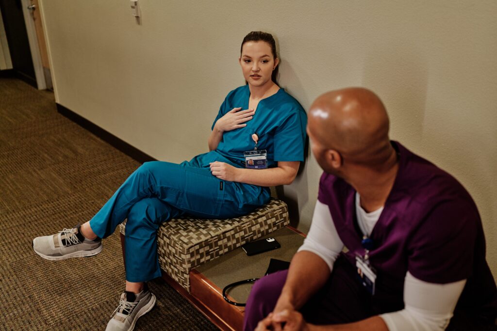 Two healthcare workers in scrubs take a break in a hospital corridor. A registered nurse in teal scrubs with an RN badge sits on a patterned bench against the wall, gesturing as she speaks, while a colleague in purple scrubs sits on the carpeted floor in the foreground, listening. A stethoscope and phone rest nearby.