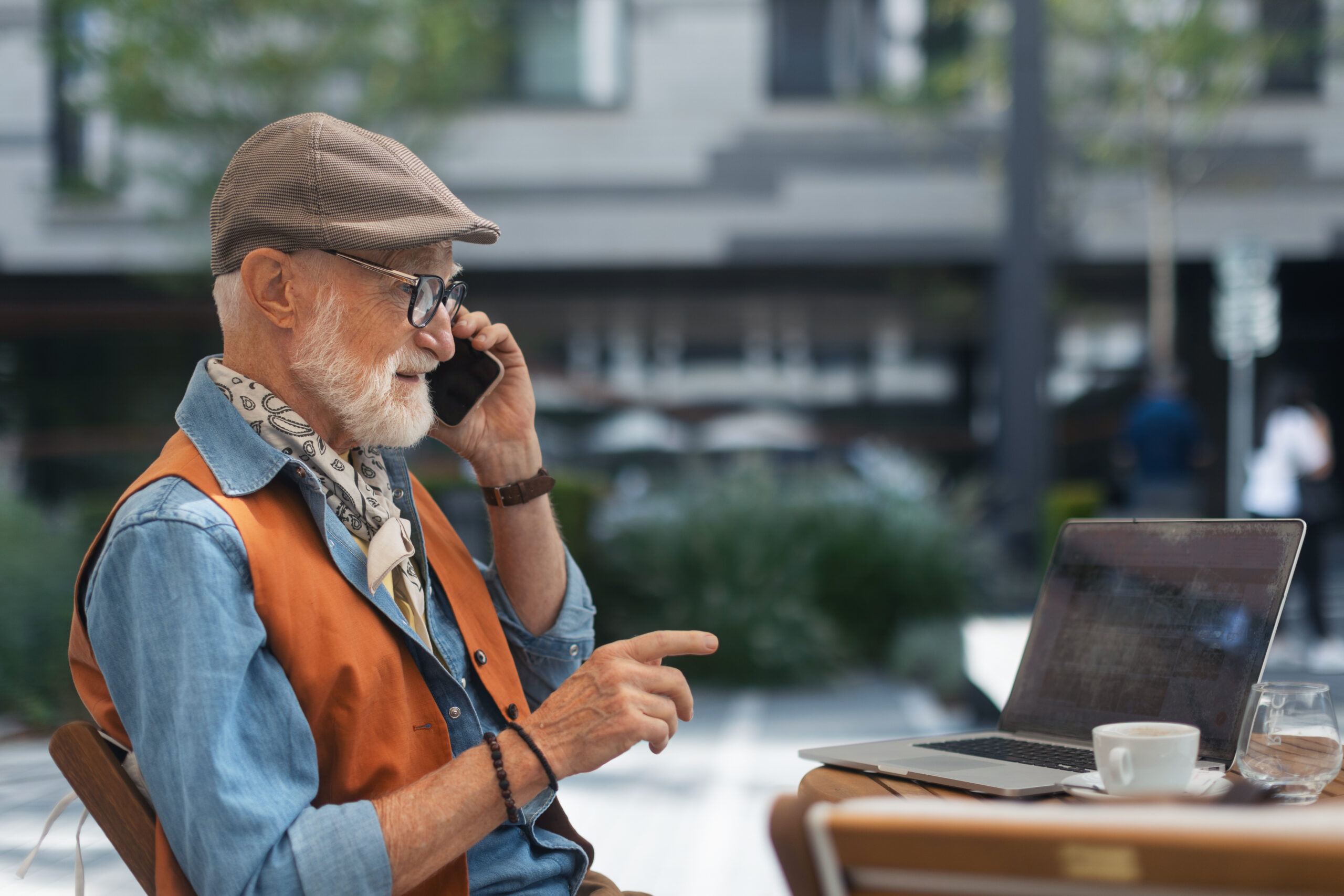 An older person with a white beard, wearing a flat cap, glasses, a denim shirt with a paisley neckerchief, and an orange vest, sits at an outdoor café table talking on a mobile phone. A laptop, coffee cup, and glass of water are on the wooden table, with a blurred urban streetscape in the background.