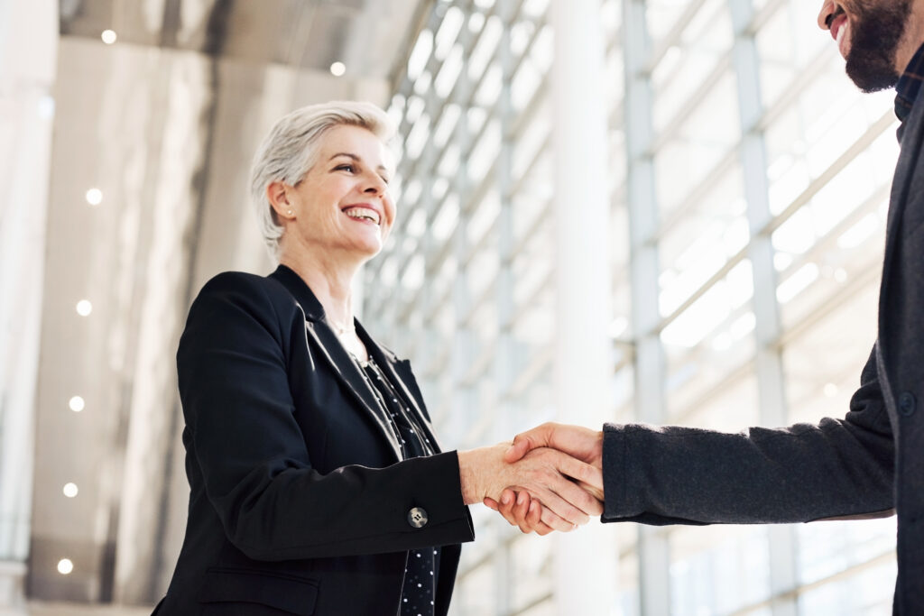 An older person with short white hair, wearing a black blazer over a polka-dot blouse, smiles while shaking hands with someone in a dark suit jacket. They stand in a bright modern building lobby with tall glass windows in the background.