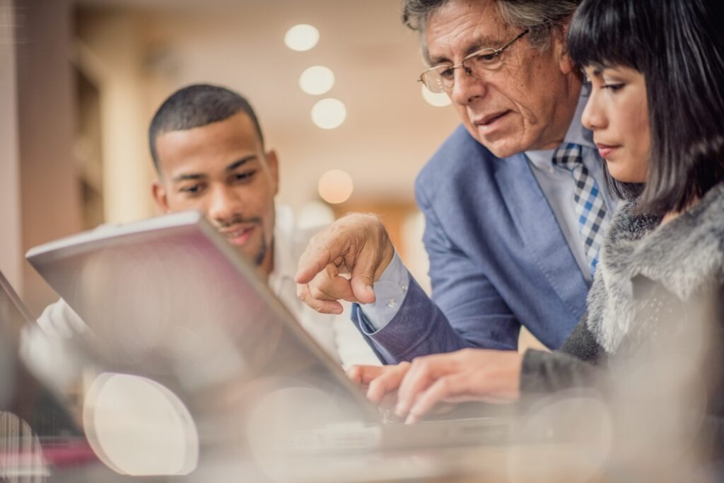 An older person in a blue button-down shirt and tie, wearing glasses, leans in and points to a laptop screen while two younger colleagues look on. The scene takes place in a warmly lit interior with soft bokeh lights in the background.