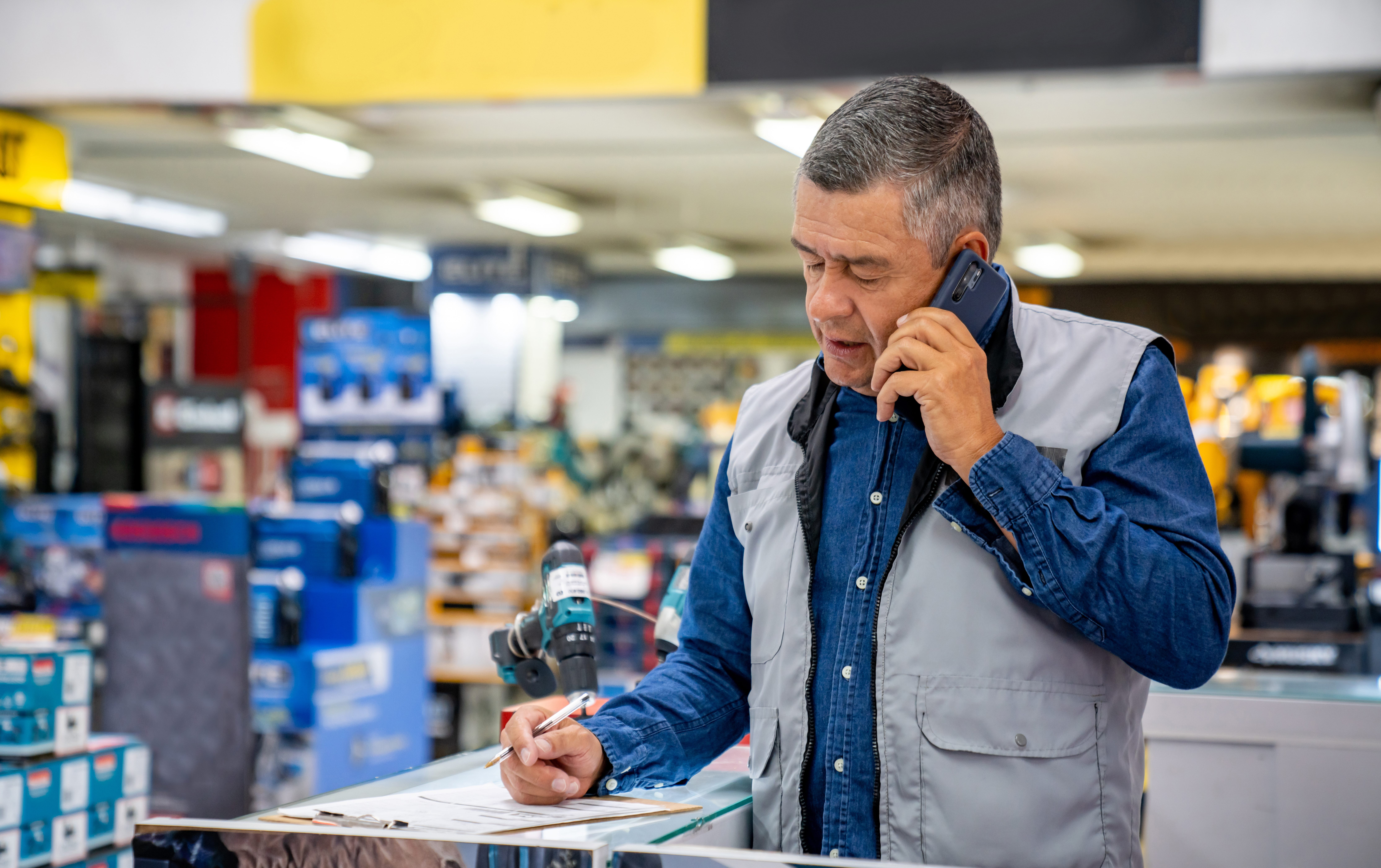 A retail worker in a gray utility vest over a blue denim shirt stands at a hardware store counter, holding a phone to their ear with one hand while writing on a clipboard with the other. Aisles of power tools and store displays are visible in the soft-focus background.