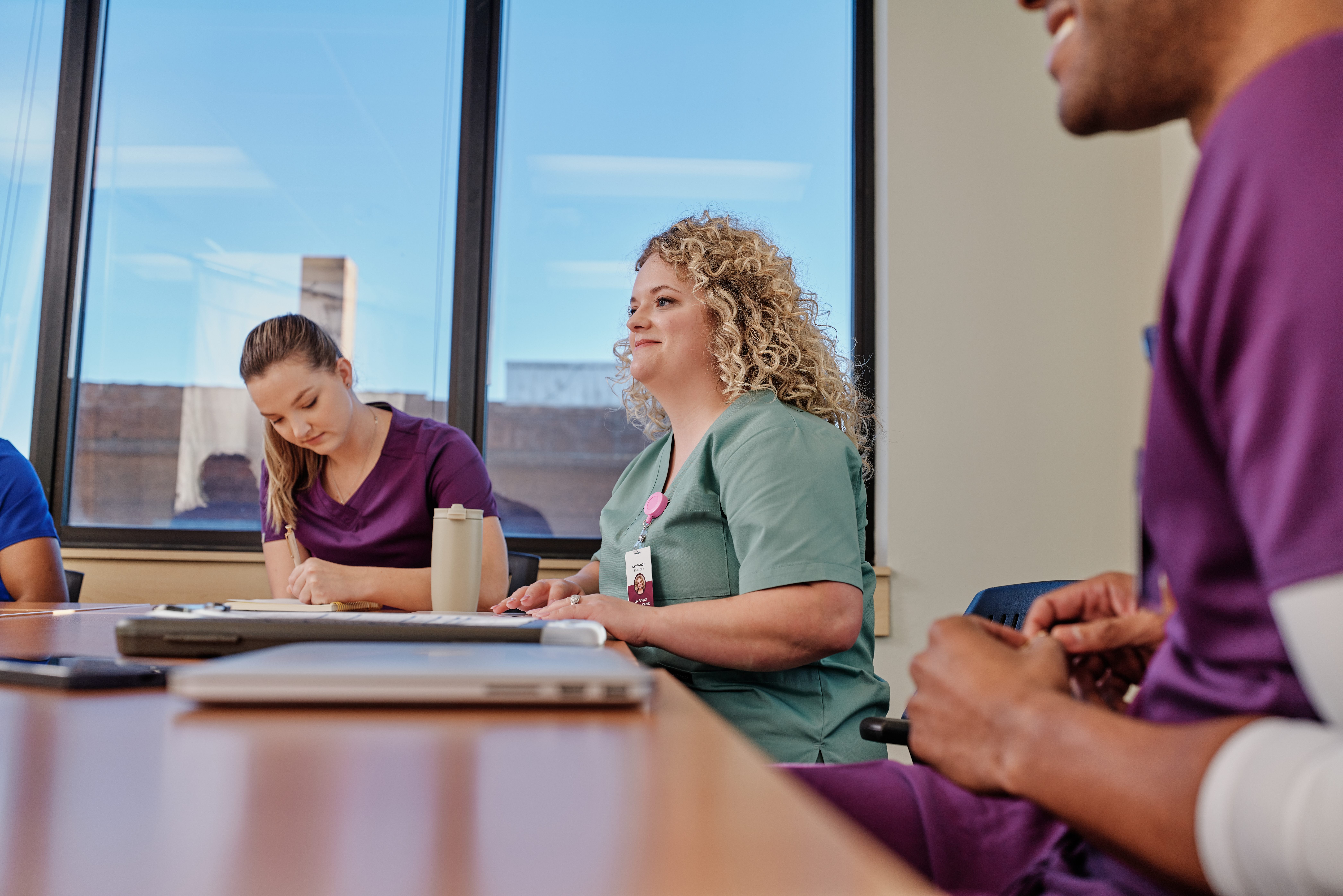 Medical staff in scrubs meeting around a boardroom table with laptops and notebooks, with one woman in green scrubs wearing an ID badge.