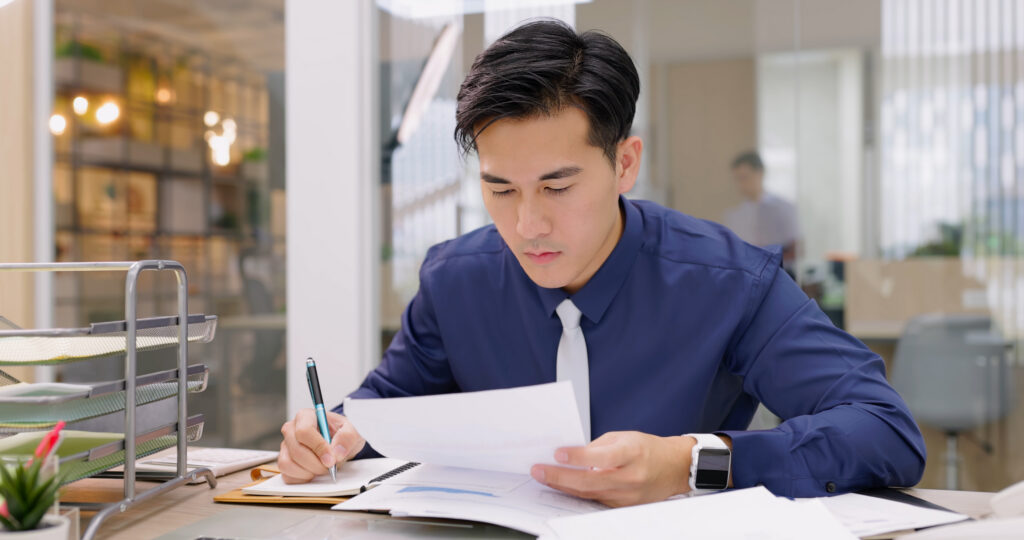 A businessman in a blue shirt and tie reviews documents with charts while writing in a notebook at his desk.