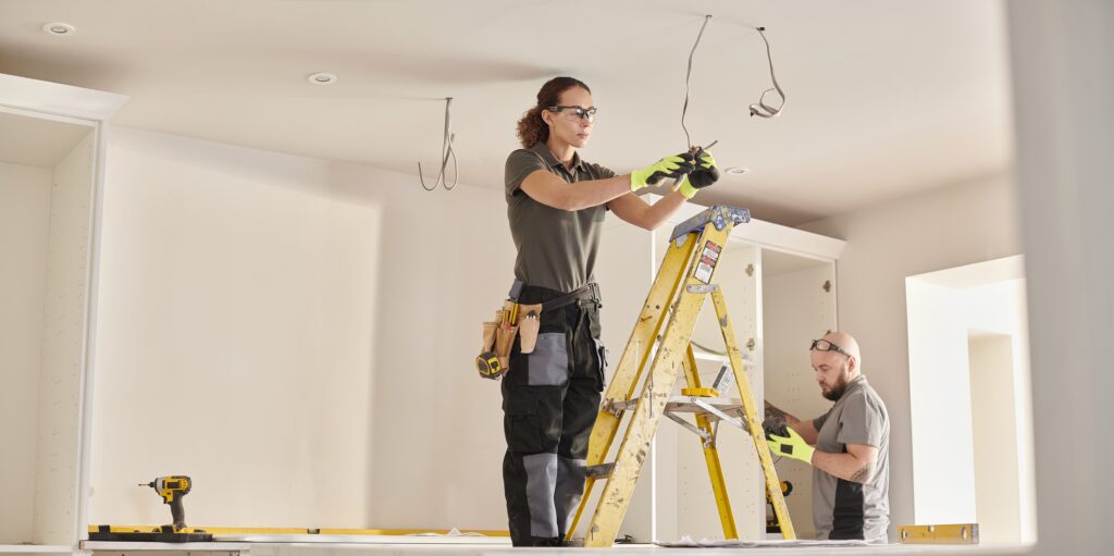A worker on a ladder wearing safety glasses and a tool belt runs electrical wiring near a ceiling, while a second worker in the background kneels to install cabinetry, with a drill and level visible on the floor.