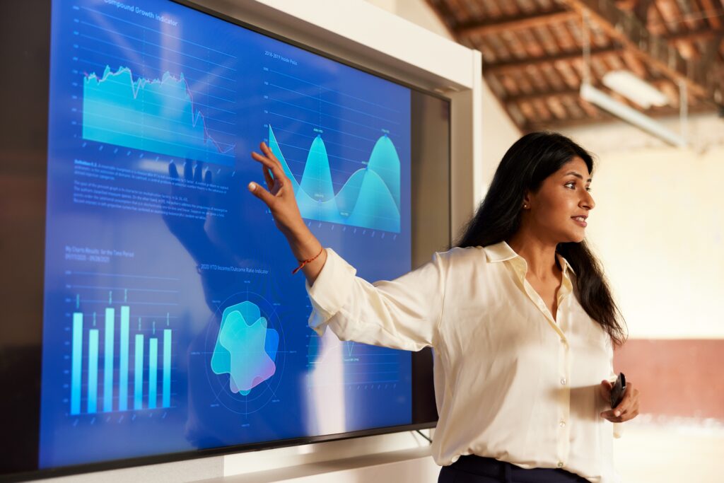 A person in a white shirt gestures toward a large screen displaying financial and growth charts, including a line graph, wave chart, bar chart, and radial data visualization.