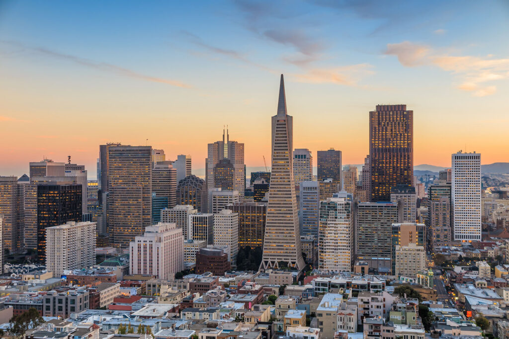 An aerial view of a downtown skyline at sunset, dominated by a pointed pyramid-shaped skyscraper among dense high-rise office buildings, with residential neighborhoods and hills visible in the distance.