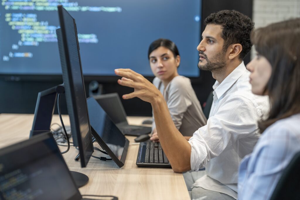 A group of colleagues at a desk with multiple monitors displaying programming code, with one person gesturing while speaking to two others seated nearby, in front of a large screen also showing code.