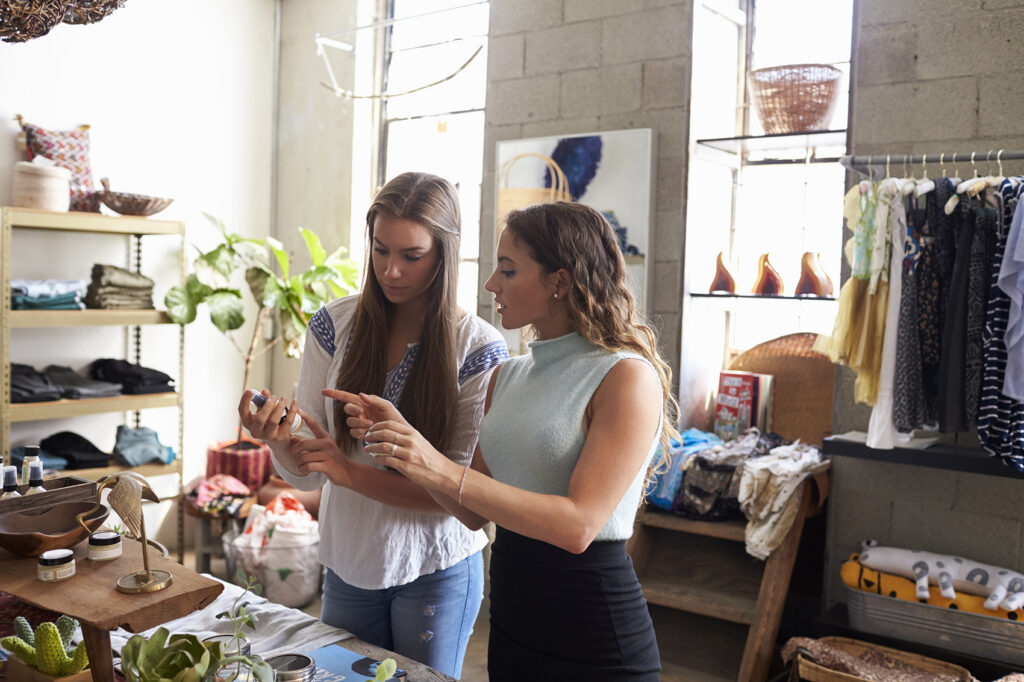 Two people stand at a counter in a retail shop, one explaining a product to the other, surrounded by clothing, home goods, and shelving.