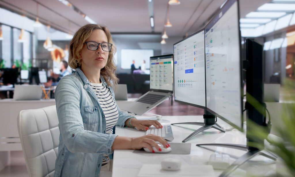A person wearing glasses works at a desk with multiple monitors displaying data dashboards and charts, with a laptop and a busy open-plan office visible in the background.