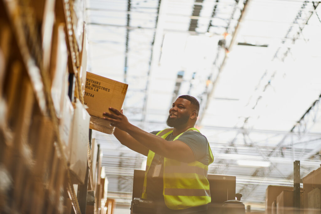 A warehouse worker in a high-visibility vest reaches up to place a cardboard box on a shelf, smiling, with warehouse racking and skylights visible above.