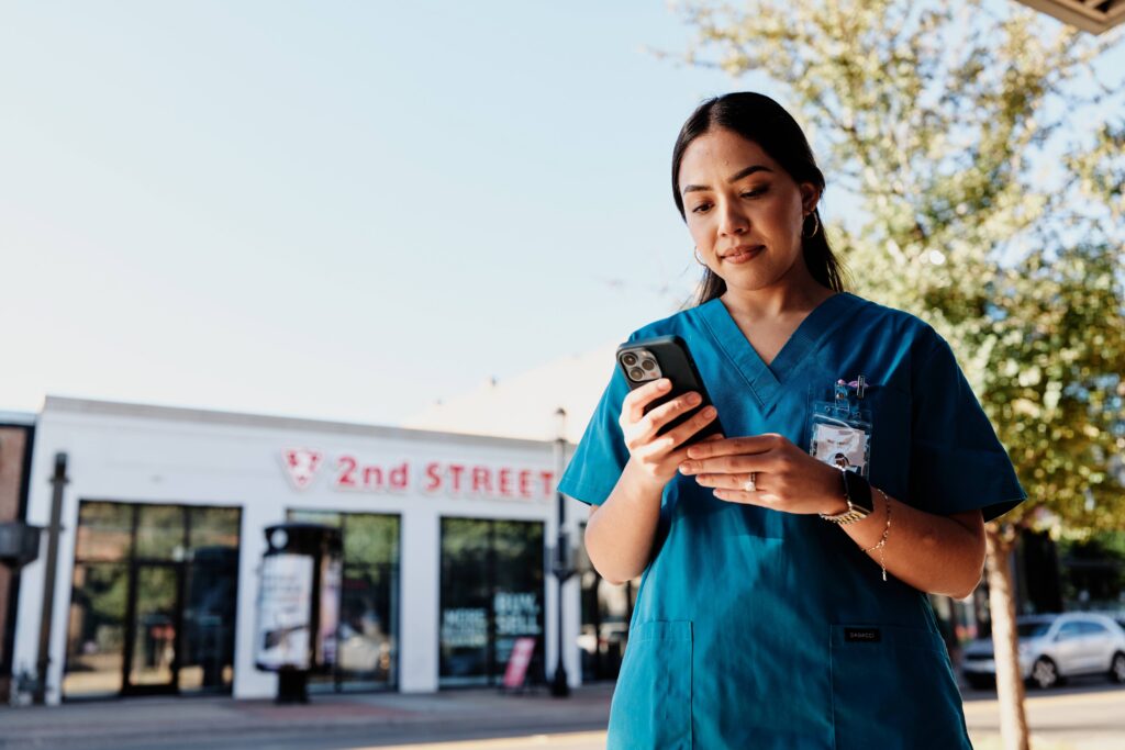 A healthcare worker in teal scrubs with a lanyard and badge looks down at a smartphone while standing on a sidewalk, with a storefront reading “2nd Street” and a tree in the background.