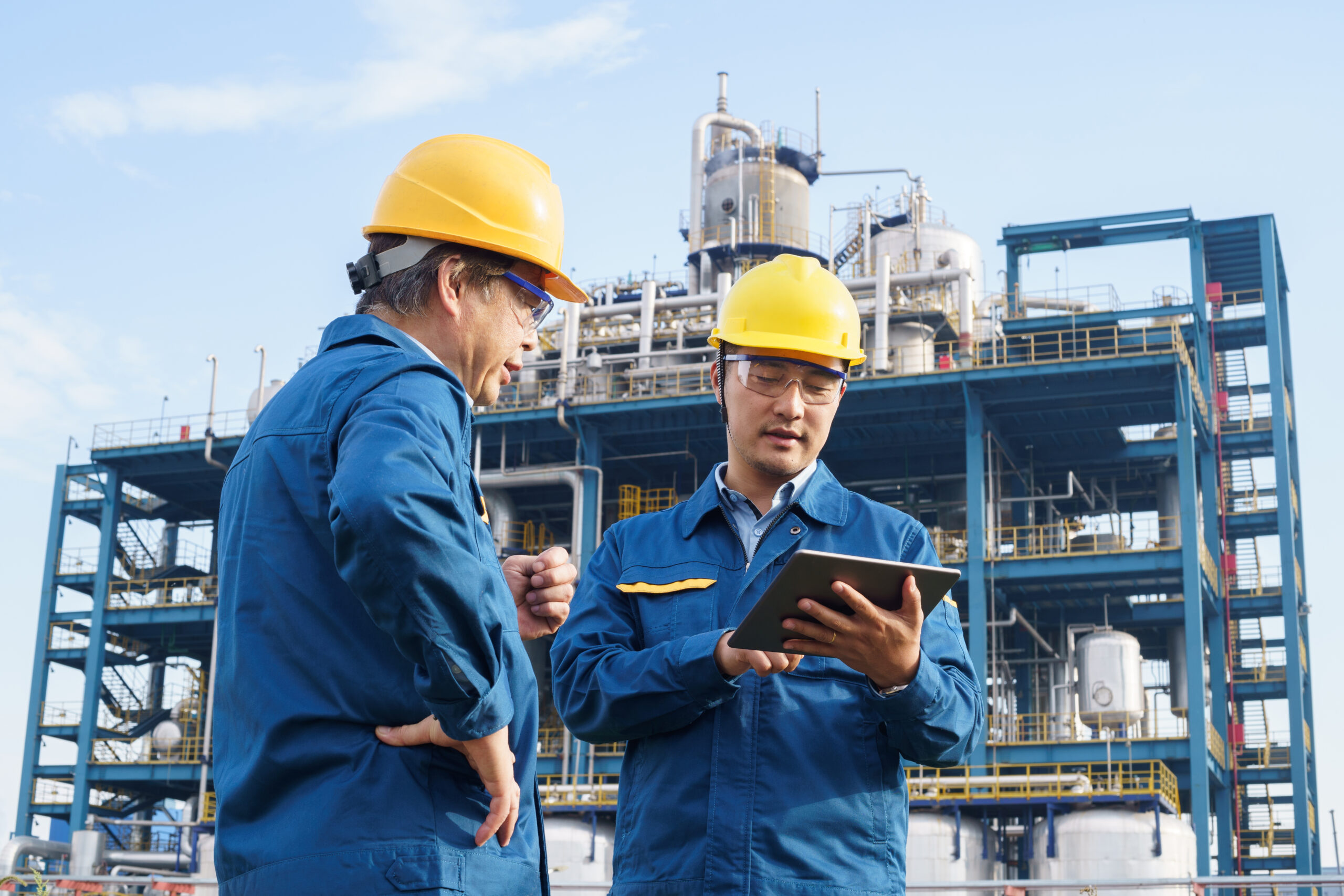 Engineers reviewing plans at a manufacturing plant, both wearing yellow hard hats with manufacturing plant in the background