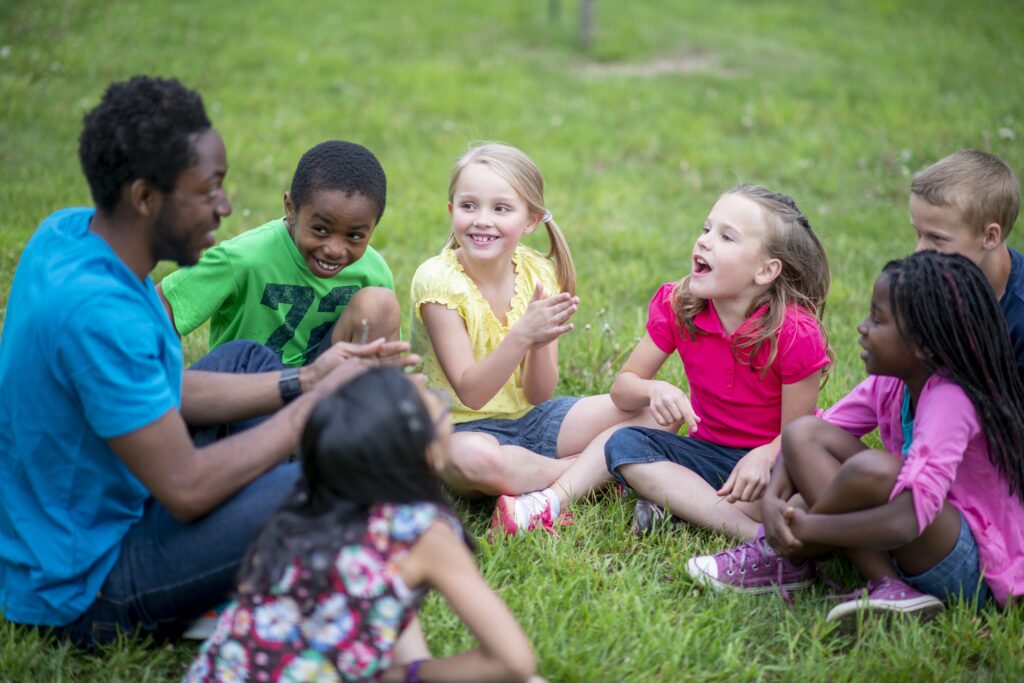 A camp counselor sits cross-legged on a grassy lawn surrounded by a diverse group of young children, all engaged in animated conversation. The children are smiling and gesturing, suggesting an interactive group activity or game.