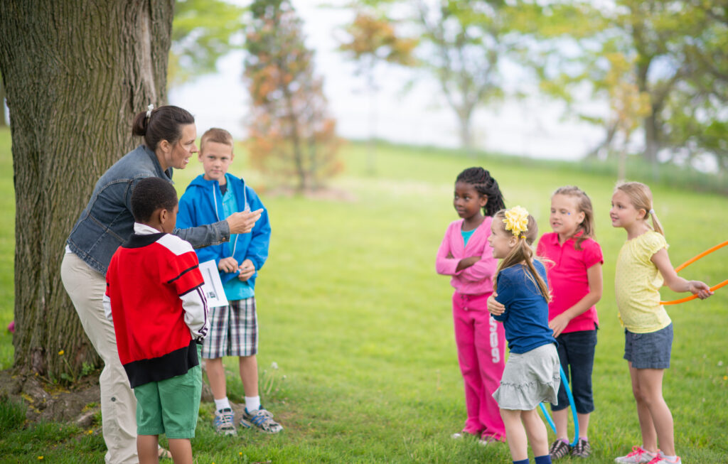 A camp counselor in a denim jacket leans down to give instructions to a group of children gathered on a grassy field beside a large tree. Two campers stand close by, while a small group of children in colorful clothing hold hula hoops a few feet away, listening attentively with more trees visible in the soft-focus background.