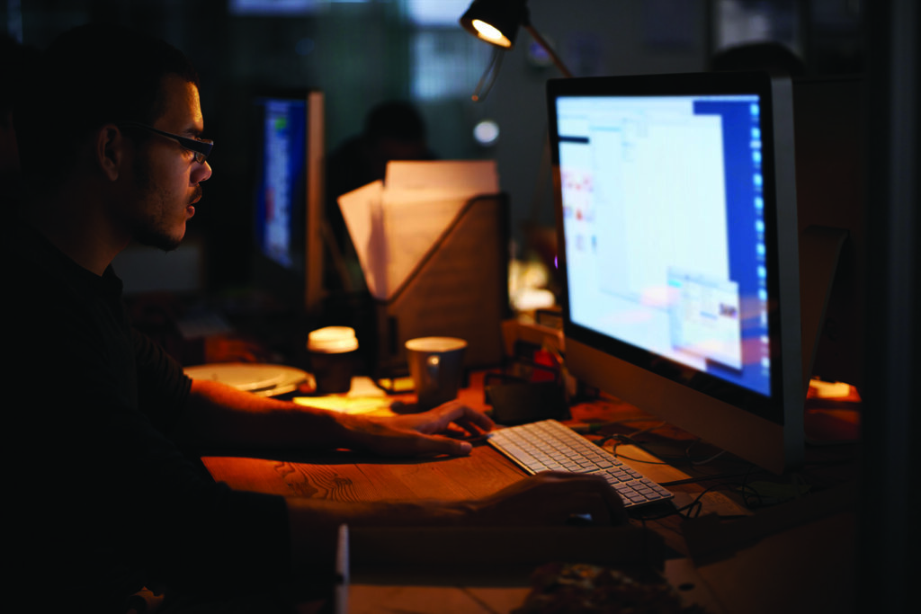 A person wearing glasses and a dark shirt sits at a wooden desk in a dimly lit office, hands resting on a keyboard in front of a large desktop monitor. A warm desk lamp illuminates coffee cups, papers, and clutter on the desk, while a second monitor and blurred figures are visible in the dark background.