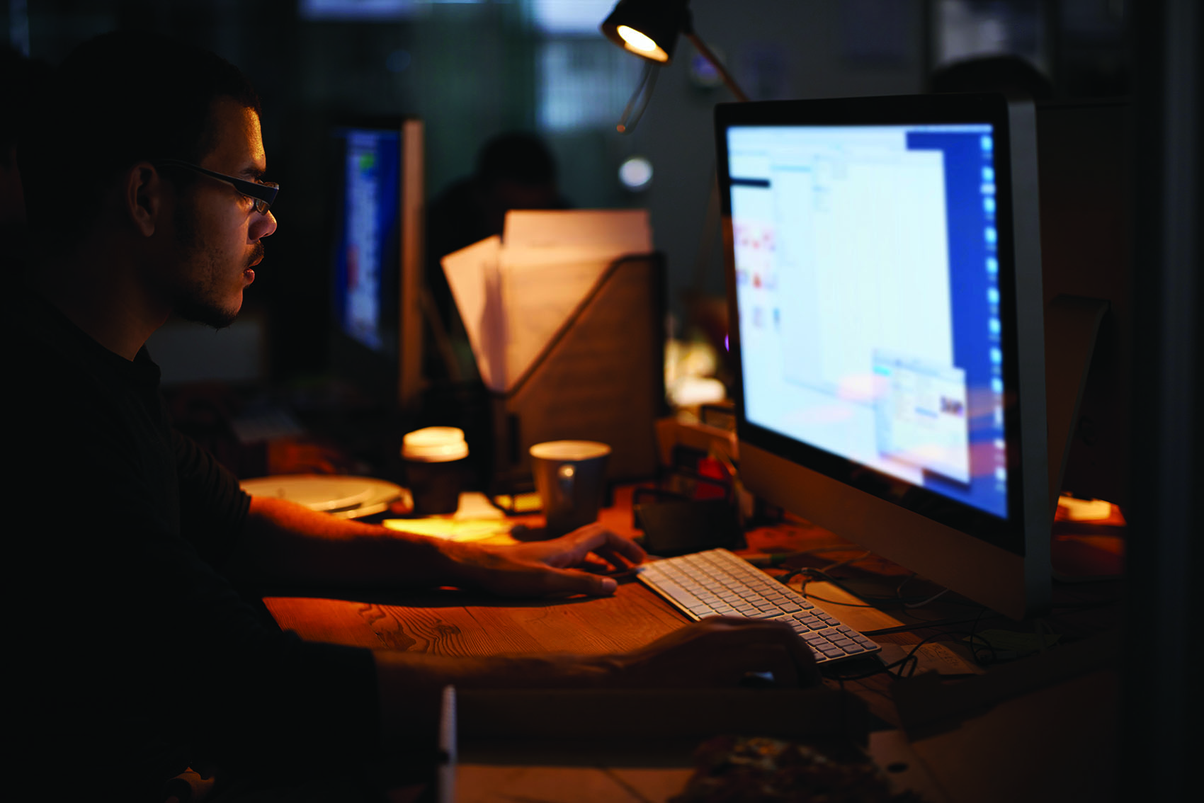 A young worker focuses on a desktop computer late into the evening, the glow of the monitor and a desk lamp lighting up an otherwise dim office.