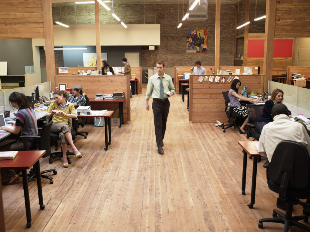 A wide view of an open-plan office with exposed brick walls, wooden beams, and hardwood floors. Several people work at desks separated by low wooden partitions — some on phones, others on laptops — while one person walks down the central aisle carrying a rolled document.