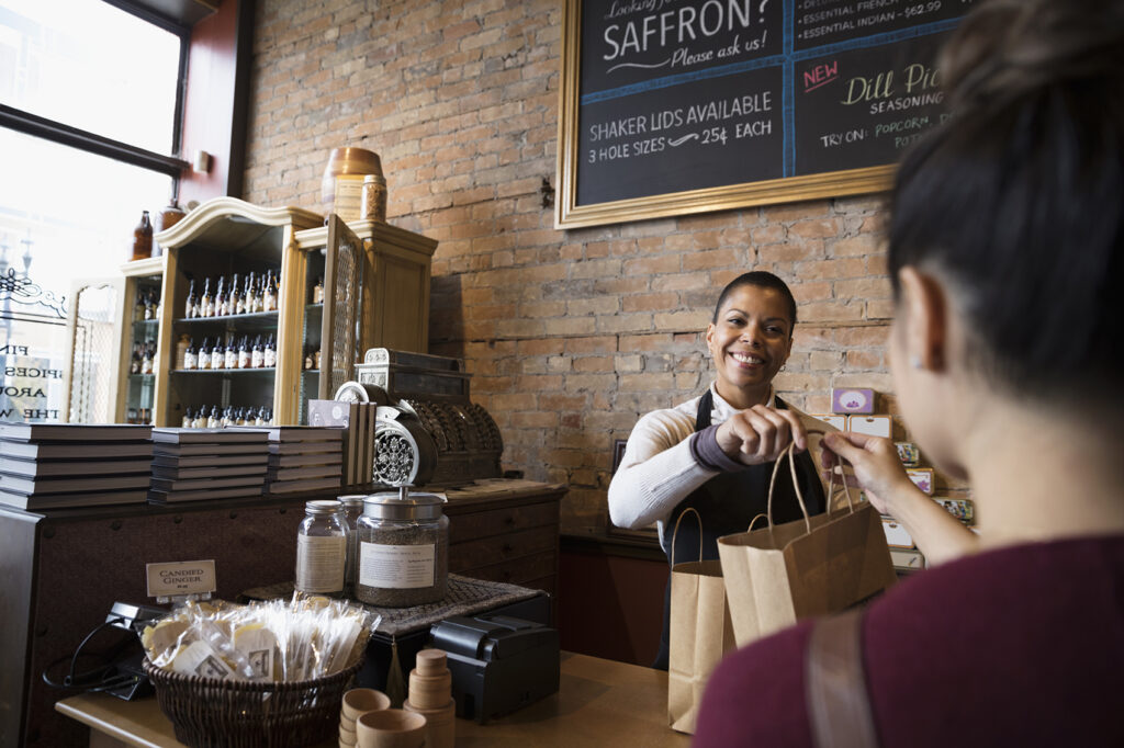 A person in a black apron stands behind the wooden counter of a small spice shop, smiling as they hand a brown paper bag across the counter to a customer with their back to the camera. Shelves of glass jars, an antique cash register, and chalkboard menus line the exposed brick walls behind them.