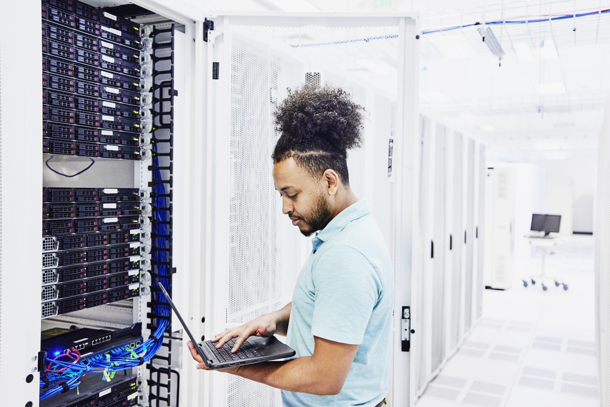 An IT professional in a server room holds an open laptop while looking at a bank of servers with blue cabling, with rows of additional server racks extending down the aisle behind him.