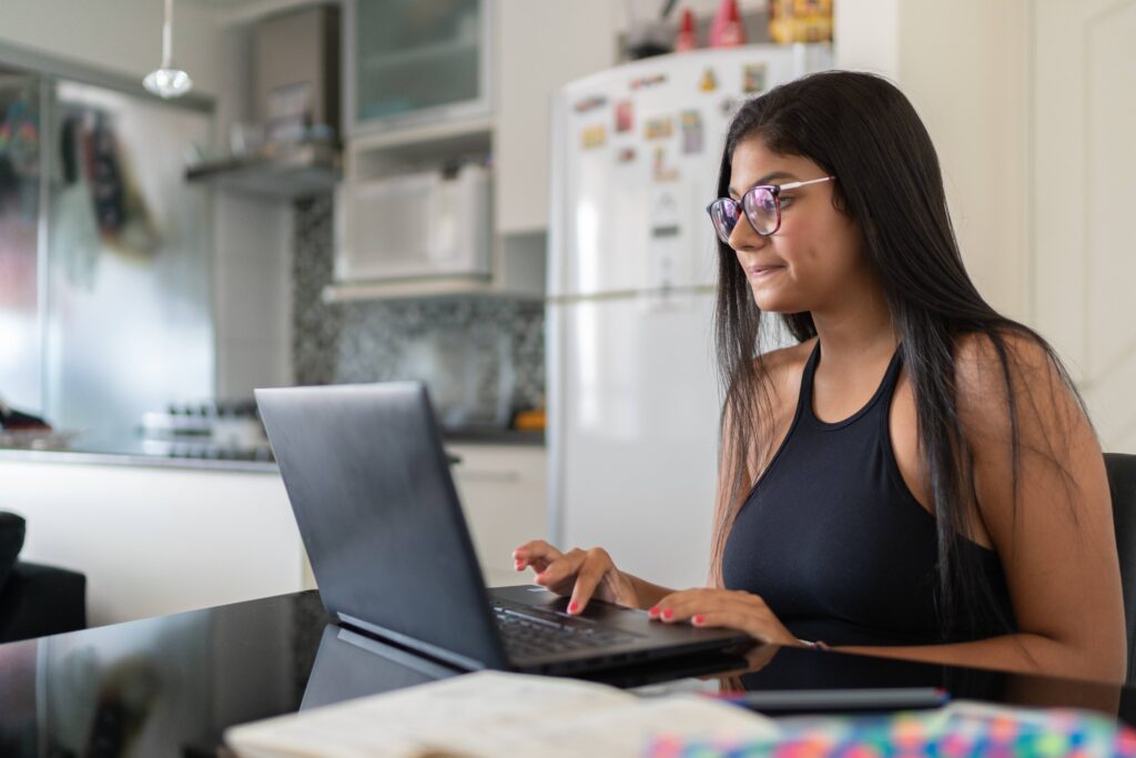 A person with long dark hair and glasses types on a laptop at a dark table in a home kitchen, with a white refrigerator covered in magnets in the background and an open notebook in the foreground.