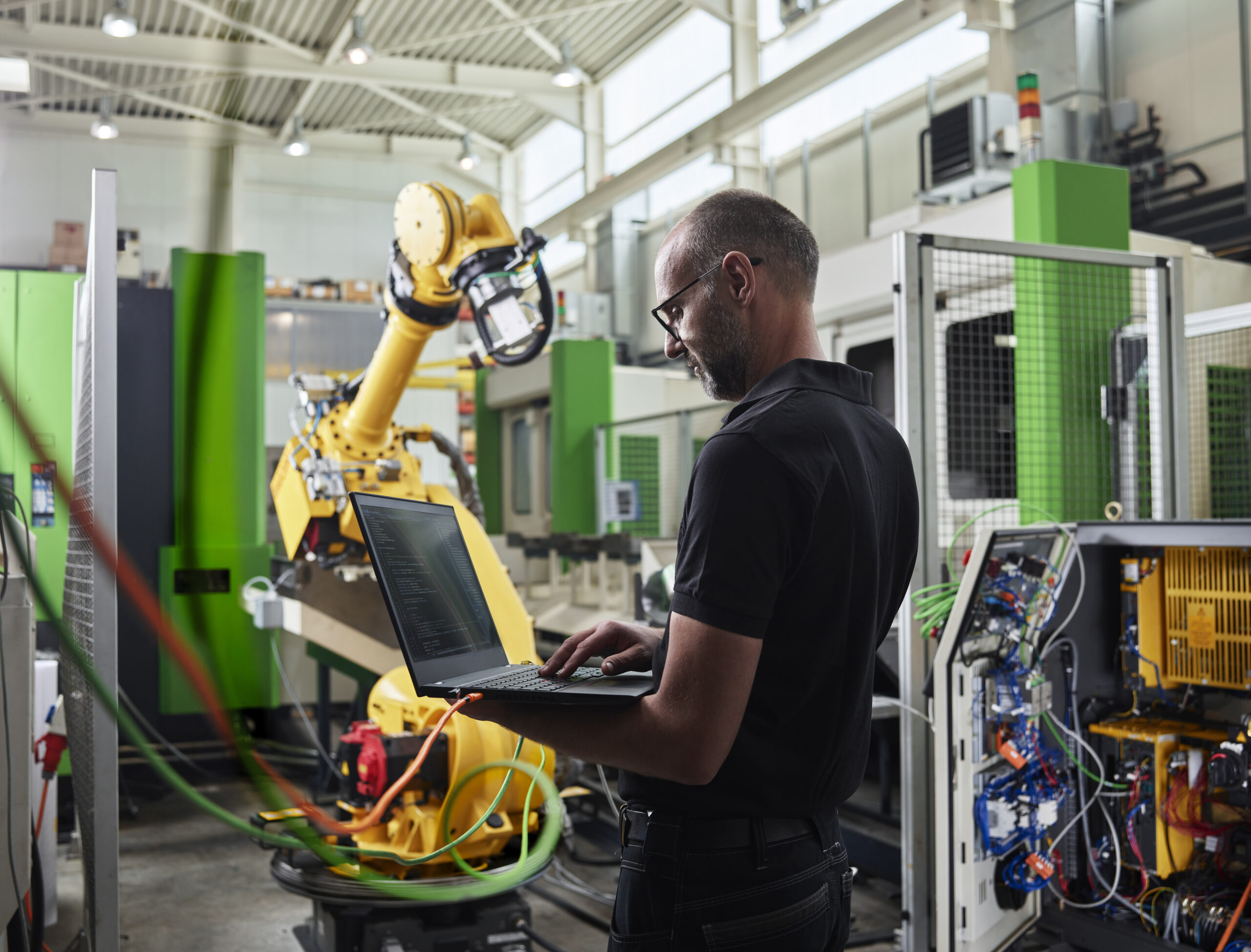 An engineer in safety glasses types on a laptop while standing beside a large yellow industrial robotic arm in a manufacturing workshop.