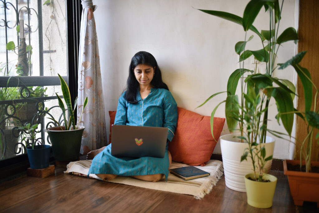 Person working on laptop sitting next to window surrounded with plant pots.