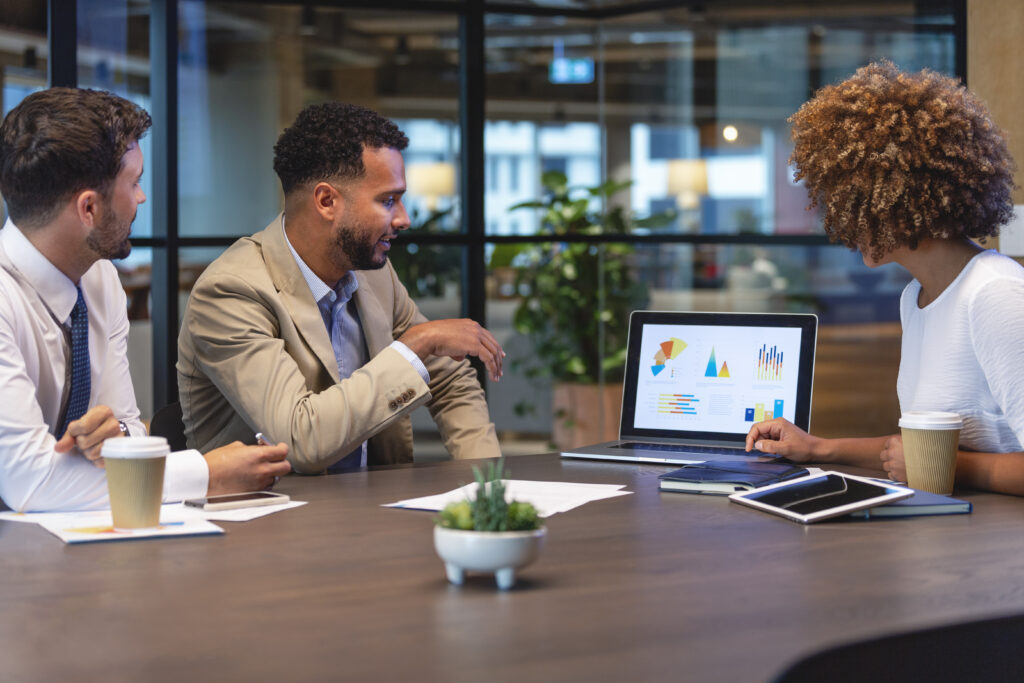 Three business people meeting and looking at a laptop. There is paperwork and other technology on the table, formal business wear. They are looking at financial graphs and charts