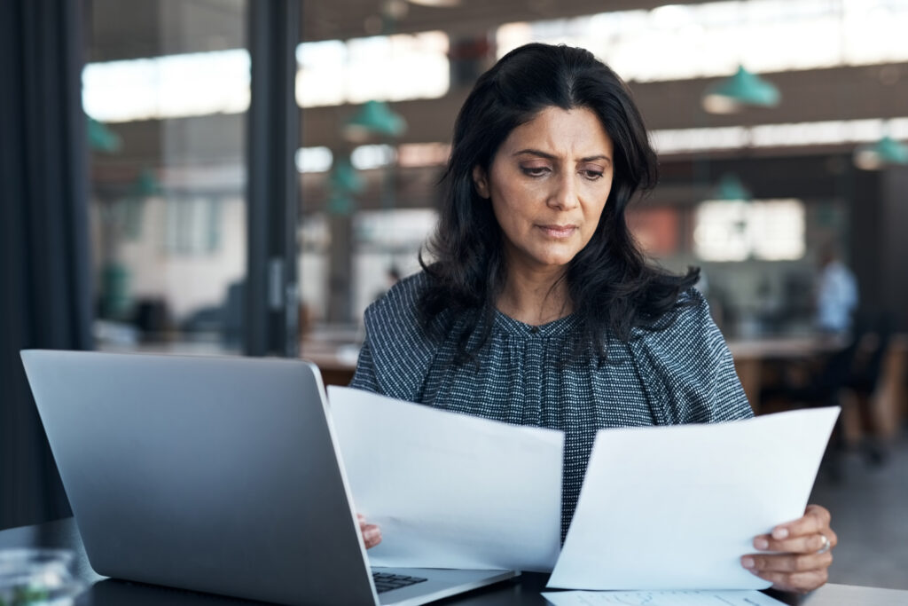 Person looking worryingly at paperwork in the office in front of their work computer with office in the background