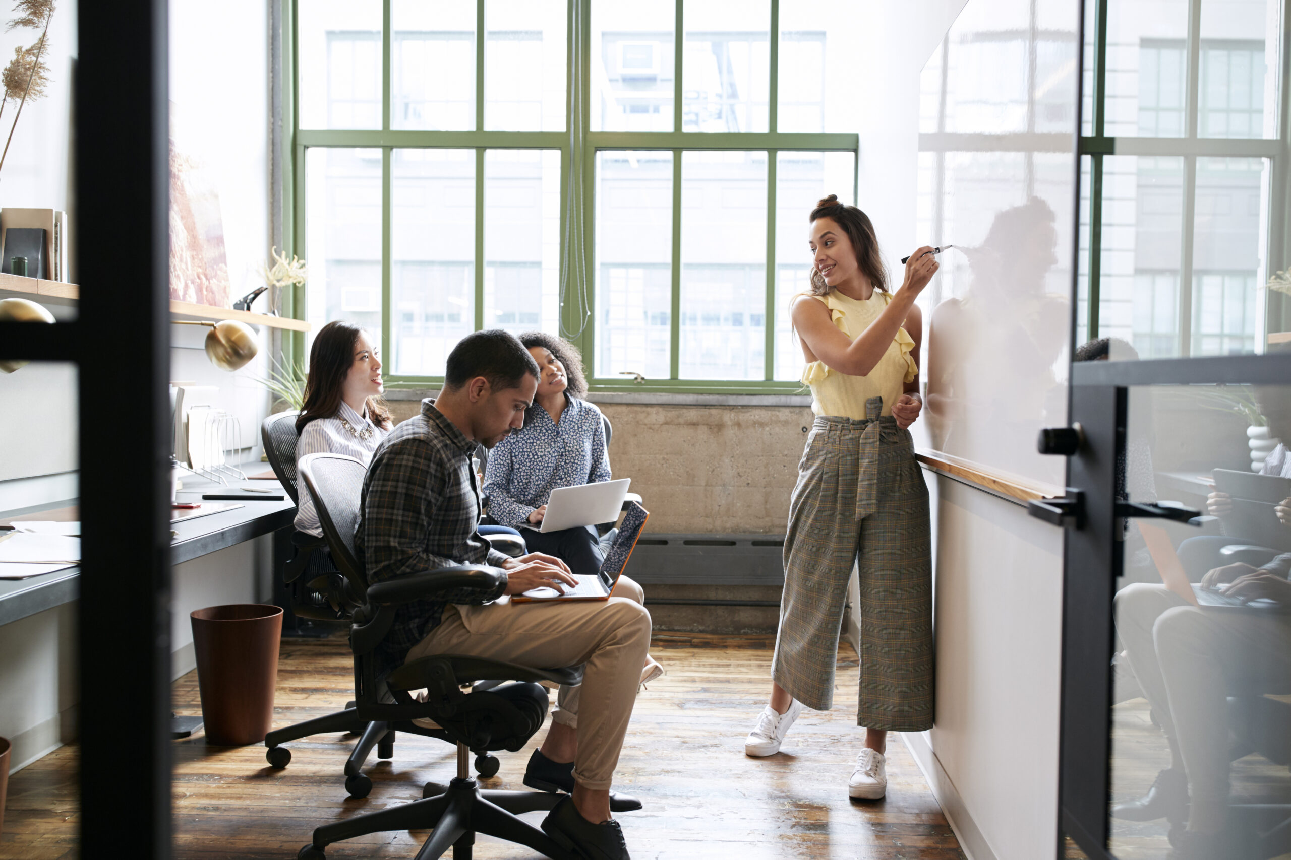 A woman in a yellow top and wide-leg plaid pants writes on a whiteboard while three coworkers — seated with laptops — look on during a collaborative meeting in a bright, modern office space with large industrial windows.