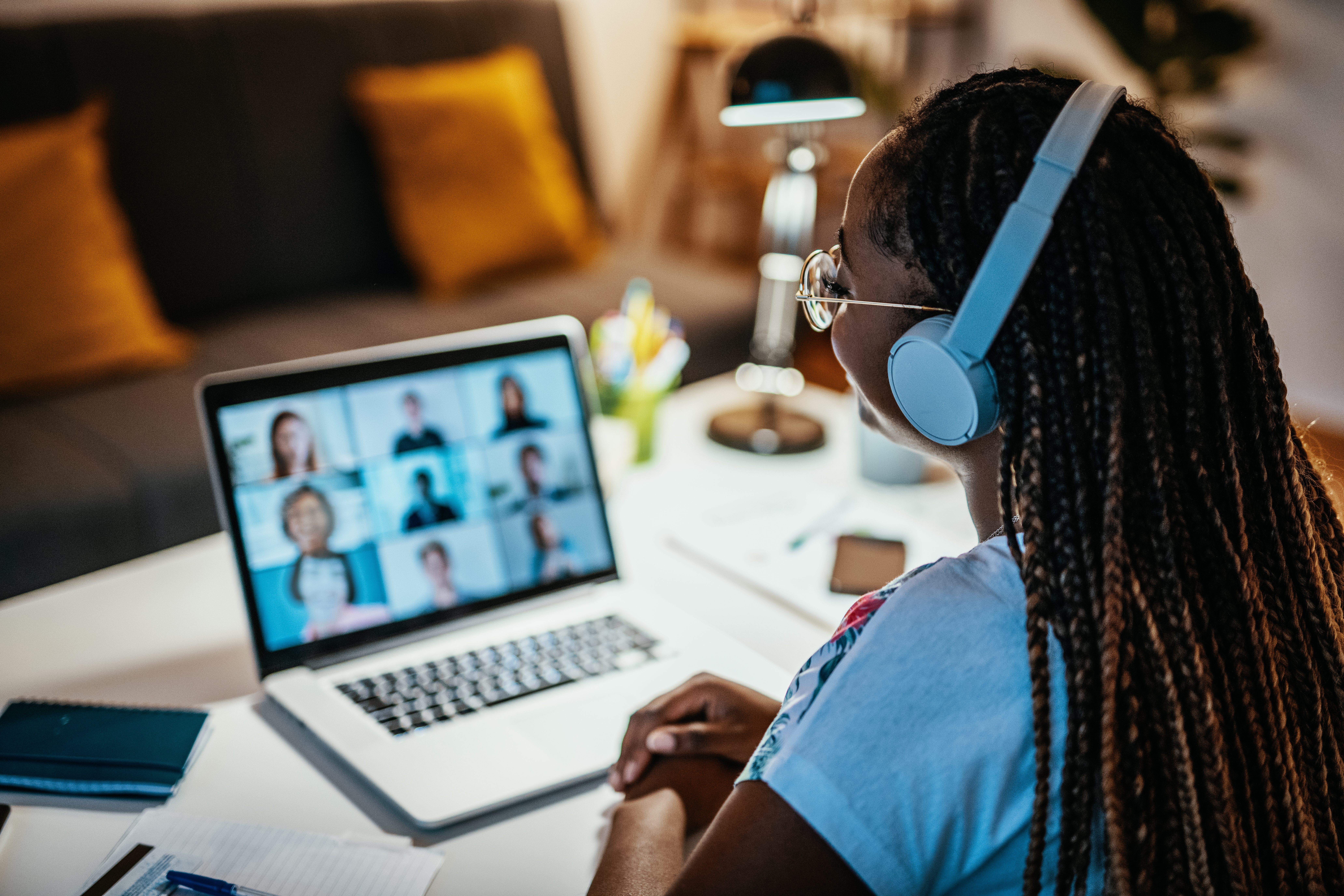 A person with long braided hair, wearing a blue T-shirt, glasses, and light blue over-ear headphones, sits at a desk facing a laptop displaying a video conference grid of nine participants. A notebook, papers, and a small plant sit on the white desk, with a couch and warm lamp light visible in the soft-focus background.