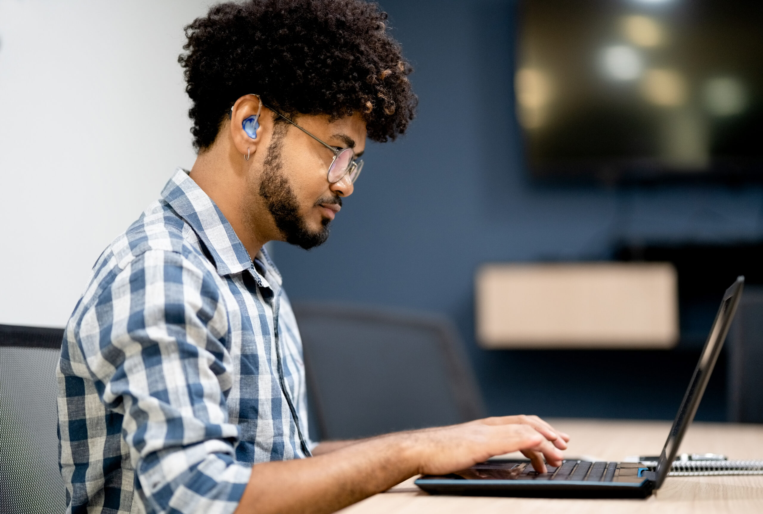 A young person with curly hair, glasses, and a hearing aid, wearing a blue and white checked shirt, sits at a wooden desk typing on a laptop in a modern office. A notebook is open beside them, with a monitor visible in the soft-focus background.
