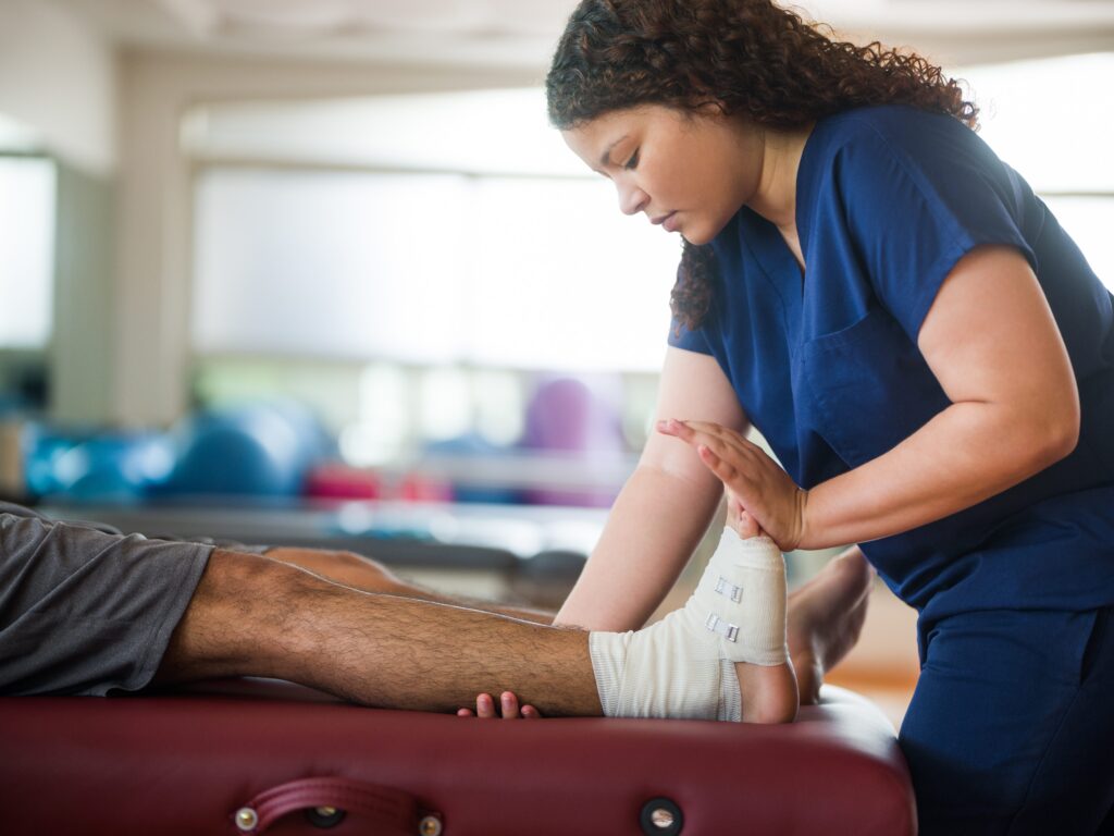 A healthcare worker in blue scrubs treats a patient's bandaged foot and ankle while the patient lies on a treatment table, with exercise balls and rehabilitation equipment visible in the background.