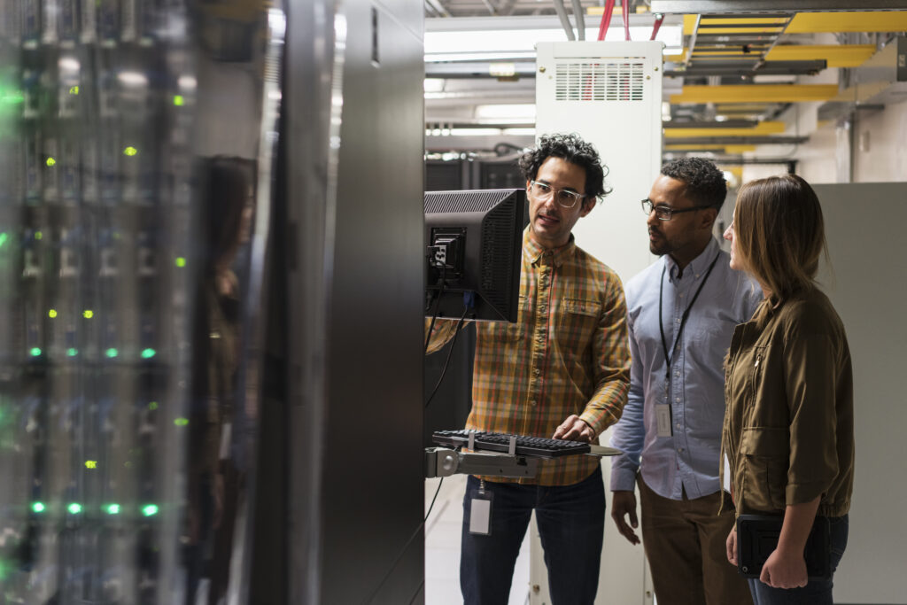 Three colleagues stand in a server room, with one person gesturing toward a monitor mounted on server equipment while explaining something to the other two, surrounded by server racks and overhead cable trays.