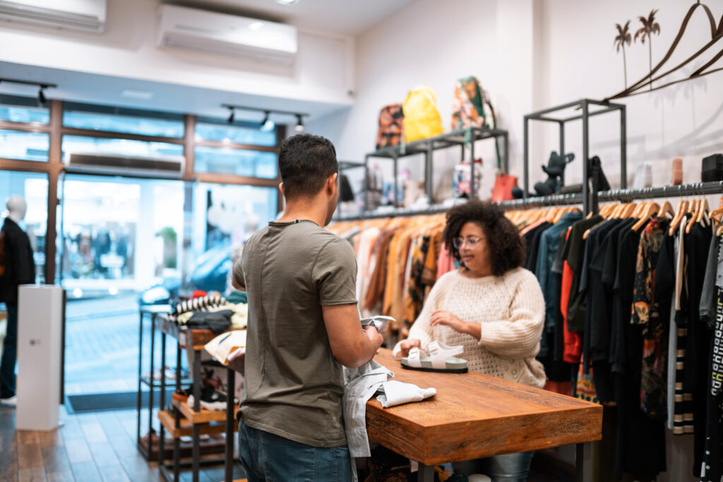 A shop assistant attends to a customer at a wooden table in a clothing store, with sandals and folded garments between them and clothing racks in the background.