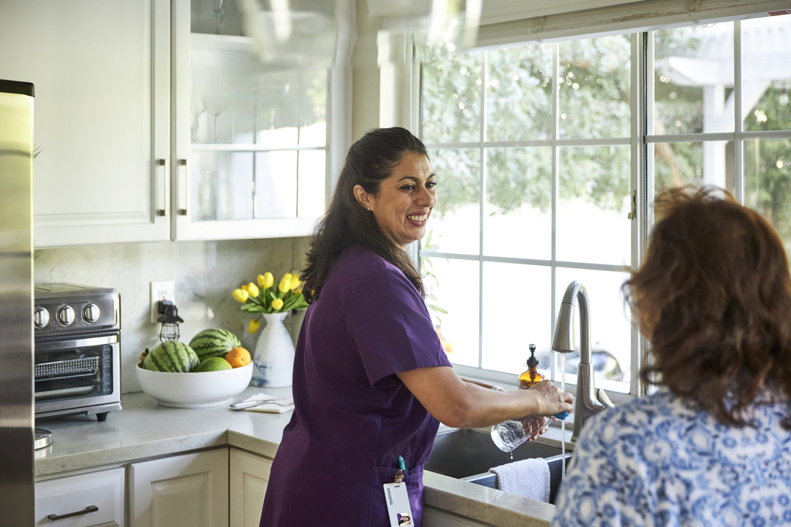 Un aidant familial fait la vaisselle dans la cuisine, sourit et parle à une autre personne.