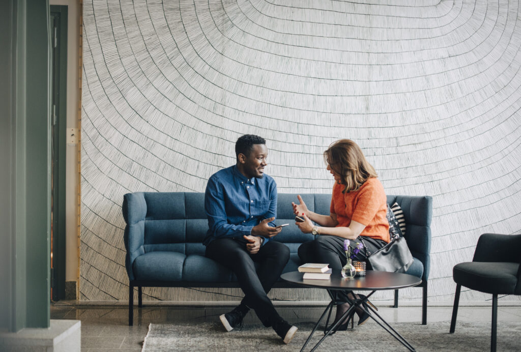 Deux personnes assises sur un canapé bleu dans un bureau moderne, engagées dans une conversation animée. Une table basse avec des livres et un vase de fleurs est visible au premier plan, devant un mur décoratif aux motifs géométriques.