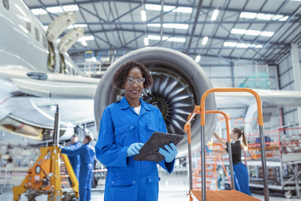 Une personne en combinaison de travail bleue, portant des lunettes de protection et des gants, tient une tablette devant un grand moteur d'avion dans un hangar aéronautique. D'autres techniciens travaillent en arrière-plan autour de l'appareil.