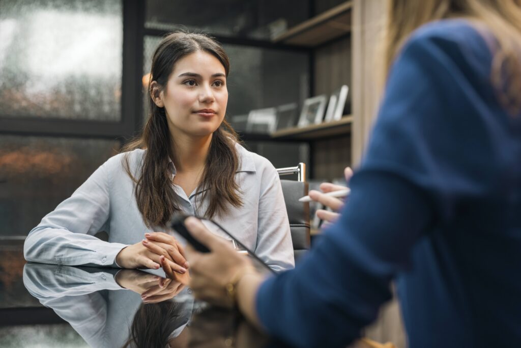 Une personne aux cheveux longs est assise à une table réfléchissante, les mains jointes, écoutant attentivement une autre personne assise en face d'elle qui tient un téléphone et gesticule en parlant.