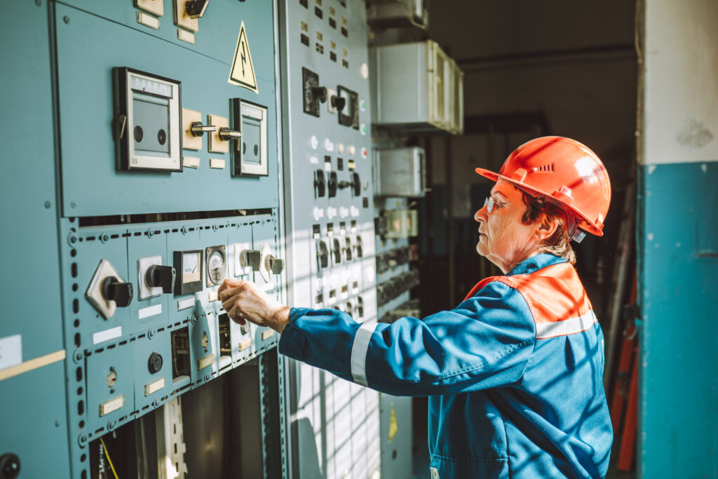 Eine Person mit orangefarbenem Schutzhelm und blau-oranger Arbeitskleidung bedient einen Schalter an einer großen industriellen Schalttafel mit zahlreichen Messinstrumenten, Reglern und Anzeigen.