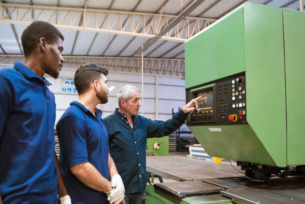 Eine erfahrene Person in einer Produktionshalle zeigt auf ein Bedienfeld einer großen grünen Maschine und erklärt es zwei jüngeren Personen in einheitlichen Poloshirts, die in einer Industriehalle stehen.