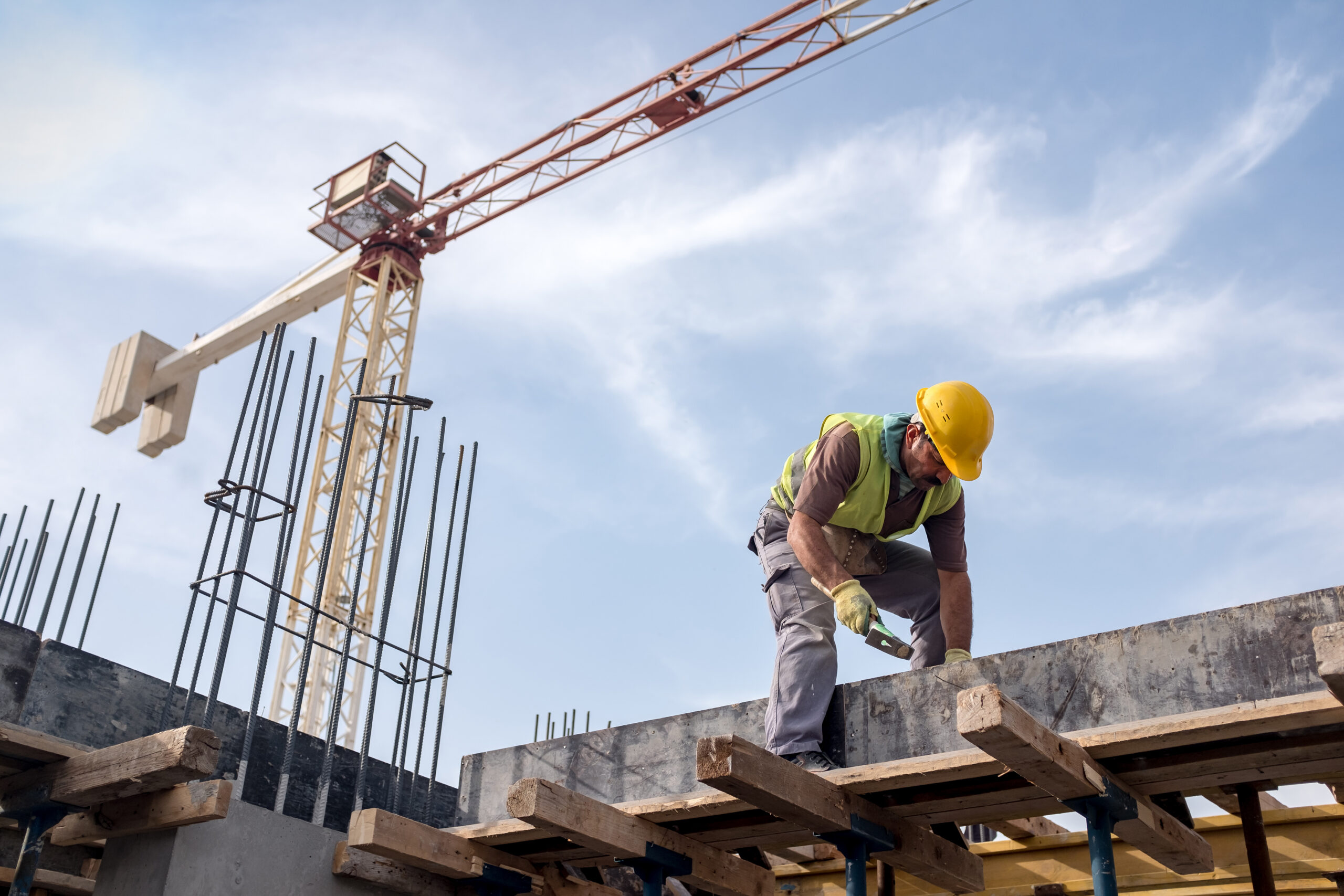 Ein Bauarbeiter mit gelbem Schutzhelm und Warnweste hämmert auf einen Betonträger auf einer Baustelle, im Hintergrund ist ein Turmkran vor dem Himmel zu sehen.