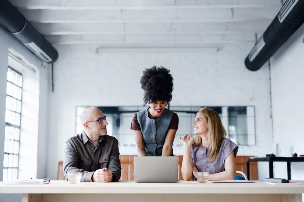 Three professionals — two seated and one standing — review content on a laptop together at a table in a bright, open office. All three are smiling and engaged in conversation.