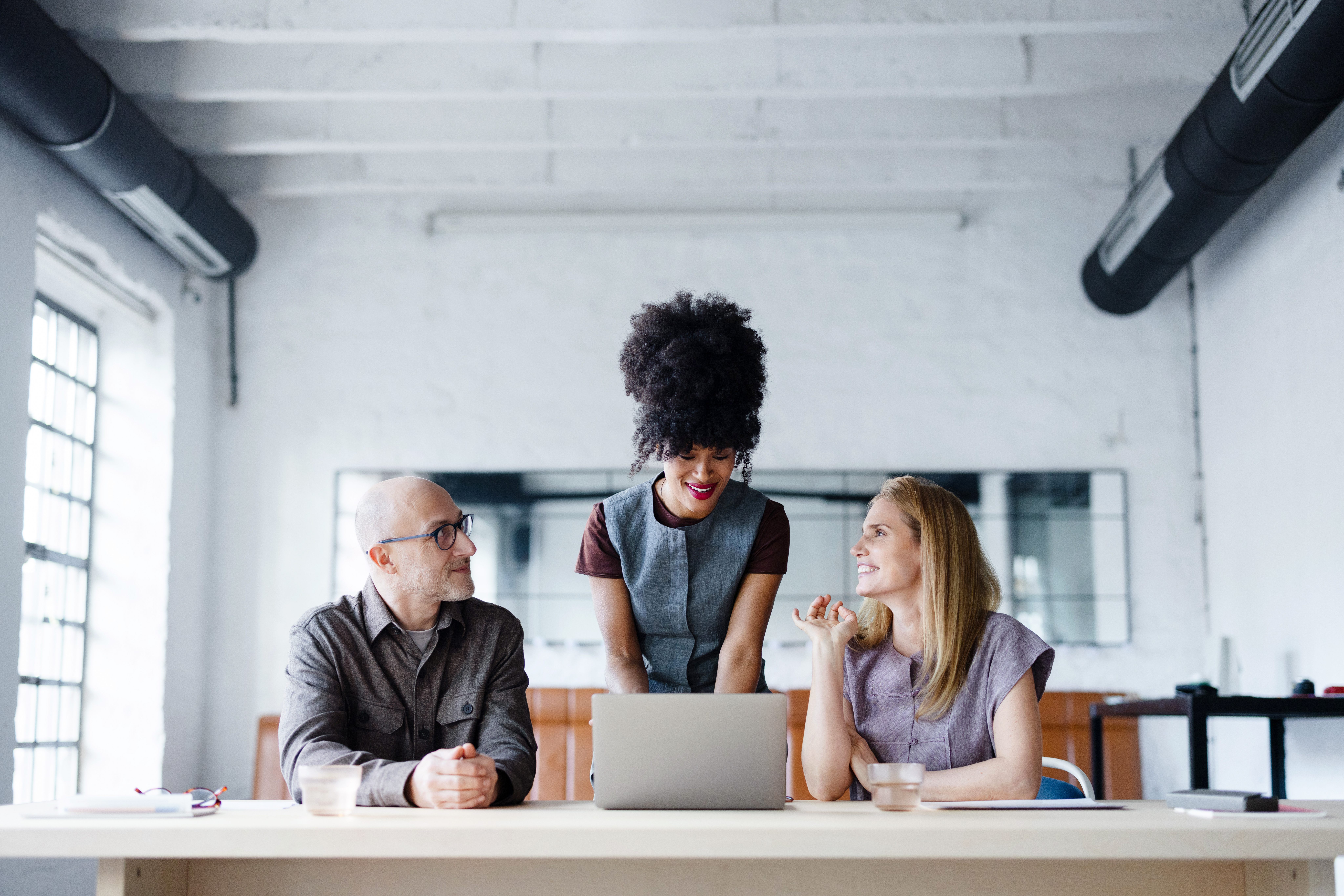 Three professionals — two seated and one standing — review content on a laptop together at a table in a bright, open office. All three are smiling and engaged in conversation.