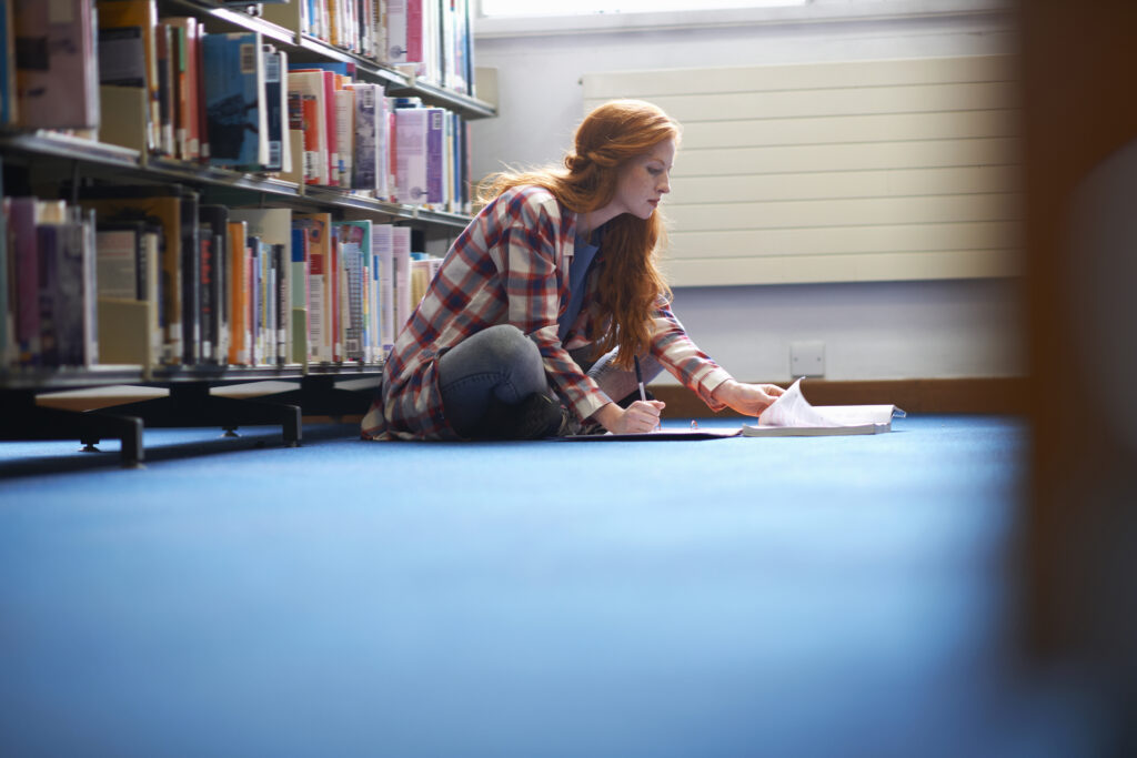 A student with long red hair sits cross-legged on the blue carpeted floor of a library, writing notes from an open book, surrounded by shelves of colourful textbooks.
