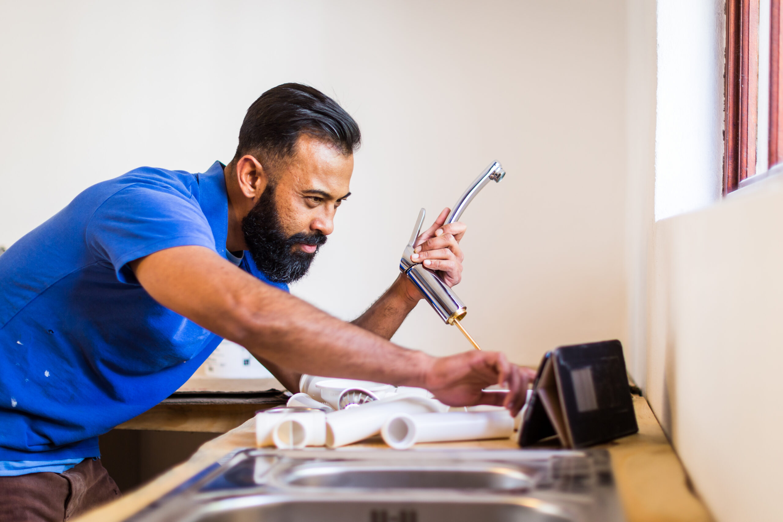 A plumber installs a new kitchen tap, consulting a tablet for reference while working alongside an assortment of pipes and fittings.