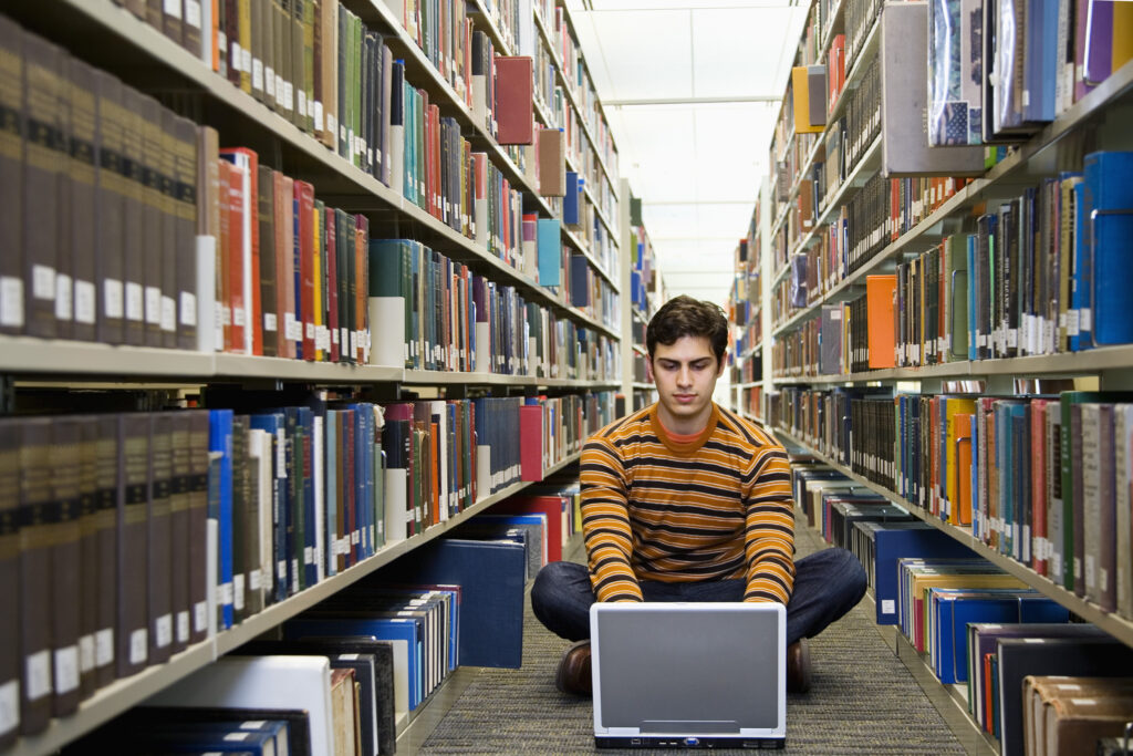 A person sits cross-legged on the floor of a library aisle, using a laptop, surrounded by tall shelves of books.